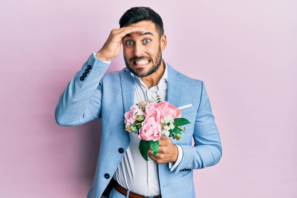 Joven empresario hispano con chaqueta de negocios sosteniendo flores estresadas y frustradas con la mano en la cabeza, cara sorprendida y enojada  - Foto, imagen