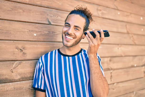 Молодой латиноамериканец улыбается счастливый слушая аудио-сообщение смартфона опираясь на стену. - Фото, изображение