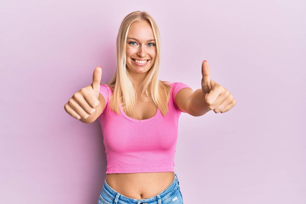 Junge blonde Mädchen in lässiger Kleidung, die positive Geste mit der Hand zustimmend, Daumen hoch lächelnd und glücklich über den Erfolg. Siegergeste.  - Foto, Bild