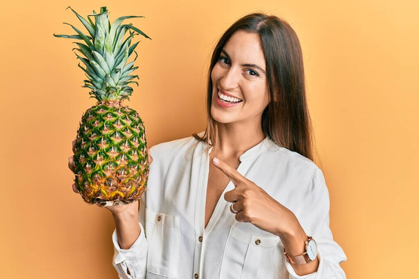 Mladá krásná žena drží ananas s úsměvem šťastný ukazující rukou a prstem  - Fotografie, Obrázek