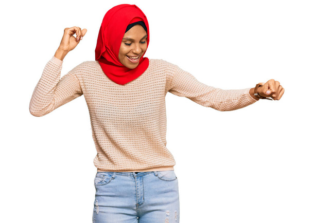 Joven mujer afroamericana vistiendo tradicional bufanda islámica hijab bailando feliz y alegre, sonriente moviéndose casual y confiado escuchando música  - Foto, imagen