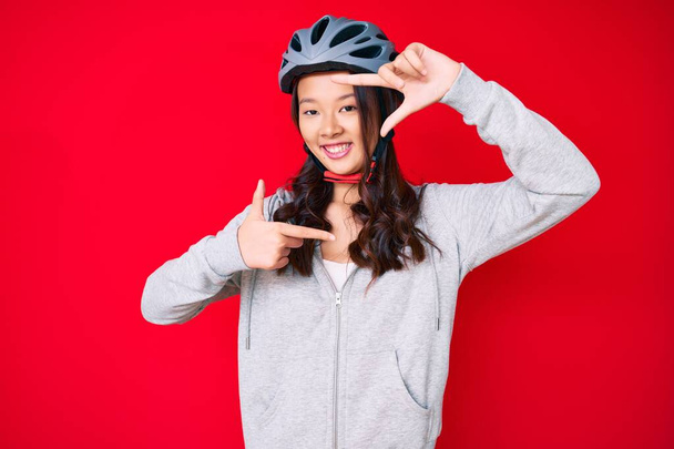 Young beautiful chinese girl wearing bike helmet smiling making frame with hands and fingers with happy face. creativity and photography concept.  - Фото, изображение