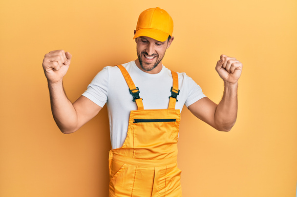 Young handsome man wearing handyman uniform over yellow background dancing happy and cheerful, smiling moving casual and confident listening to music  - Zdjęcie, obraz