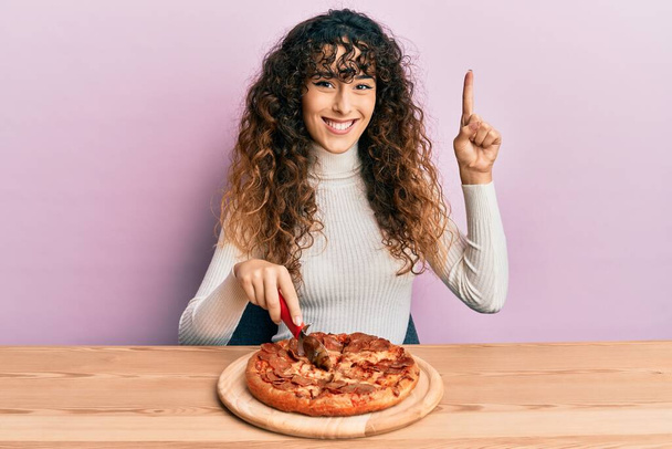 Jeune fille hispanique coupe pizza pepperoni savoureux sourire avec une idée ou une question pointant du doigt avec le visage heureux, numéro un  - Photo, image