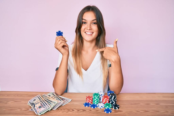 Beautiful caucasian woman playing poker holding casino chips pointing finger to one self smiling happy and proud  - Photo, Image