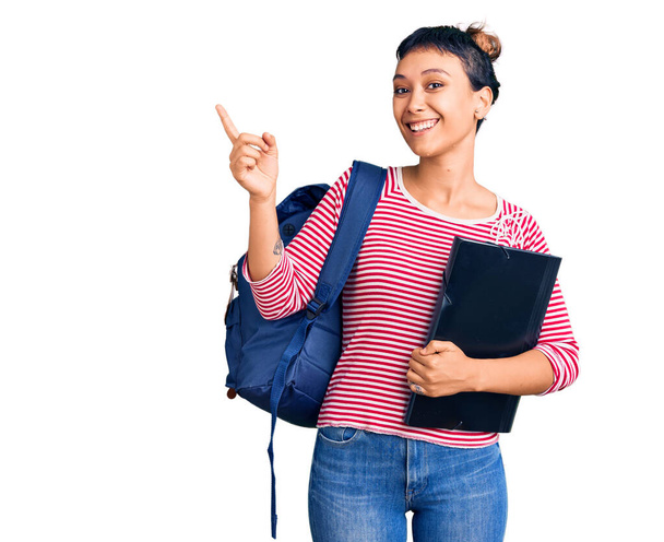 jonge vrouw dragen student rugzak houden bindmiddel glimlachen gelukkig wijzend met hand en vinger naar de zijkant  - Foto, afbeelding