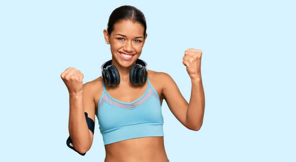 Beautiful hispanic woman wearing sportswear and headphones screaming proud, celebrating victory and success very excited with raised arms  - Fotografie, Obrázek