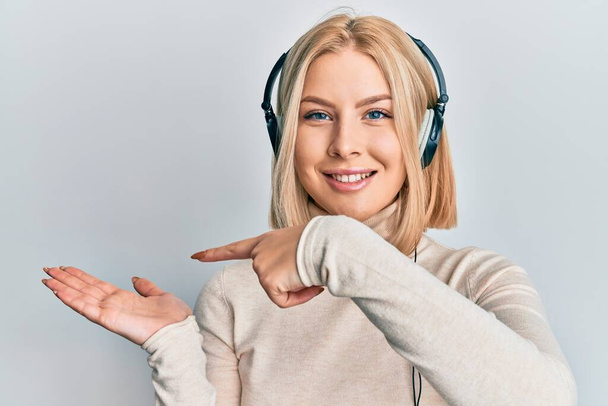 Молода блондинка слухає музику за допомогою навушників здивована і посміхається камері, представляючи рукою і вказуючи пальцем
.  - Фото, зображення