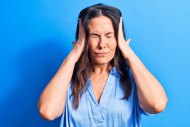 Junge schöne brünette Frau in lässigem T-Shirt vor isoliertem blauen Hintergrund mit der Hand auf dem Kopf, Kopfschmerzen wegen Stress. Migräne. - Foto, Bild