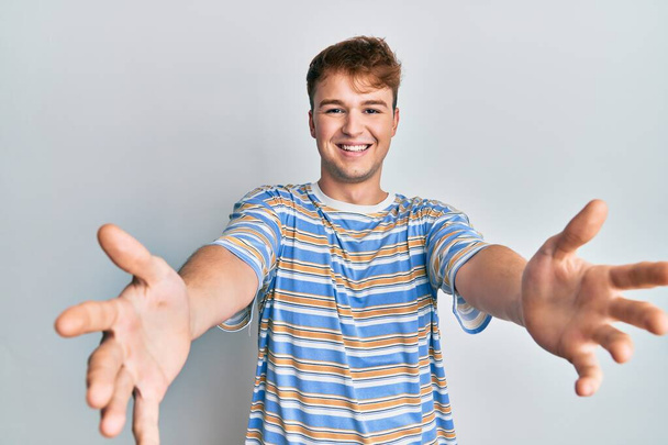 Jeune homme caucasien portant un t-shirt à rayures décontractées regardant la caméra souriant à bras ouverts pour un câlin. expression joyeuse embrassant le bonheur.  - Photo, image