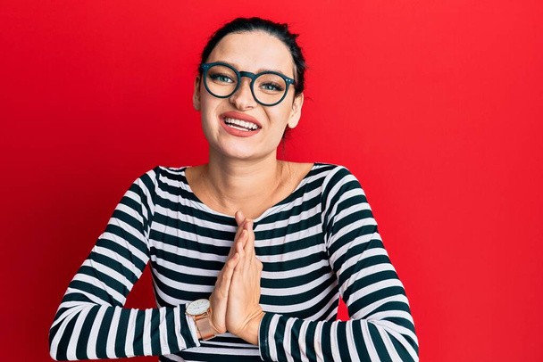 jonge Kaukasische vrouw dragen casual kleding en bril bidden met de handen samen vragen om vergeving glimlachen zelfverzekerd.  - Foto, afbeelding