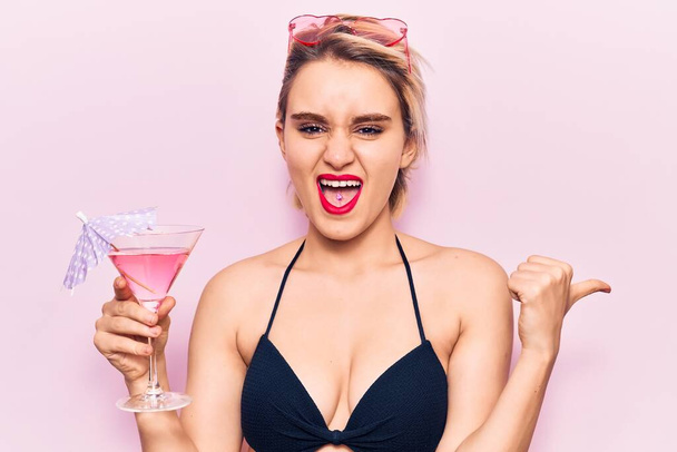 Jovem mulher loira bonita vestindo biquíni bebendo coquetel apontando o polegar para o lado sorrindo feliz com a boca aberta  - Foto, Imagem