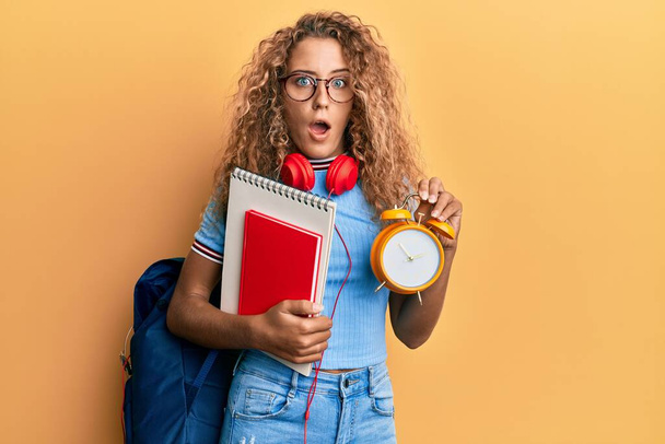 Beautiful caucasian teenager girl wearing student backpack and holding alarm clock afraid and shocked with surprise and amazed expression, fear and excited face.  - Photo, Image