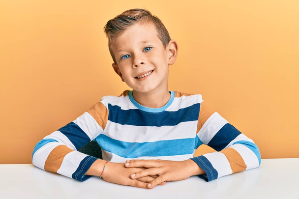 Adorable enfant caucasien portant des vêtements décontractés assis sur la table avec un sourire heureux et cool sur le visage. chanceux.  - Photo, image