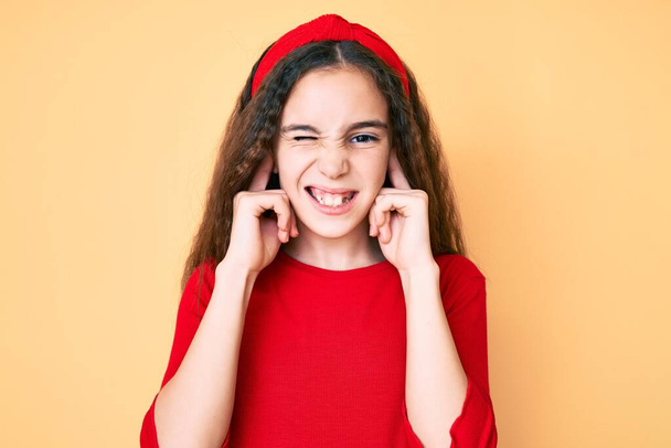 Cute hispanic child girl wearing casual clothes and diadem covering ears with fingers with annoyed expression for the noise of loud music. deaf concept.  - Foto, Imagen