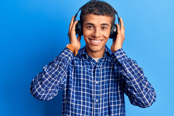Jeune homme afro-américain écoutant de la musique à l'aide d'écouteurs regardant positif et heureux debout et souriant avec un sourire confiant montrant les dents  - Photo, image