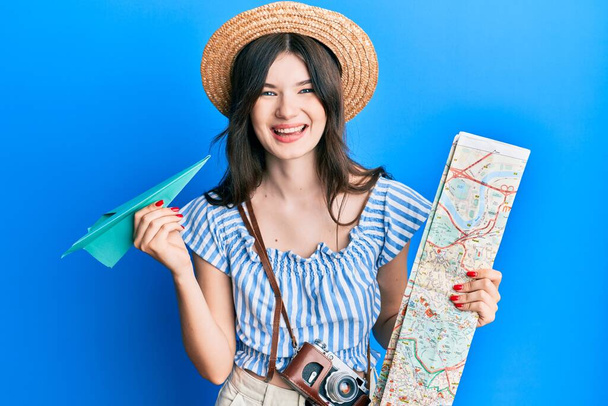Νεαρή όμορφη καυκάσια κοπέλα κρατώντας χάρτινο αεροπλάνο και χάρτη της πόλης χαμογελώντας και γελώντας δυνατά γιατί αστείο τρελό αστείο.  - Φωτογραφία, εικόνα