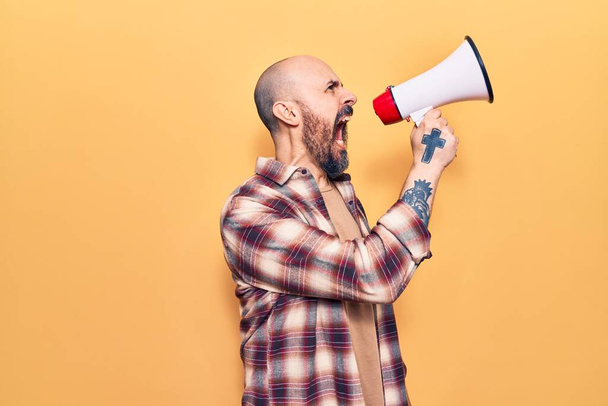 Young handsome bald man with angry expression. Screaming loud using megaphone standing over isolated yellow background - Photo, Image