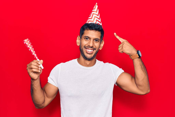 Young latin man wearing birthday hat smiling happy pointing with hand and finger  - Photo, Image
