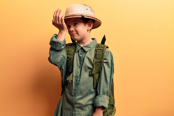 Cute blond kid wearing explorer hat and backpack surprised with hand on head for mistake, remember error. forgot, bad memory concept.  - Foto, Imagen