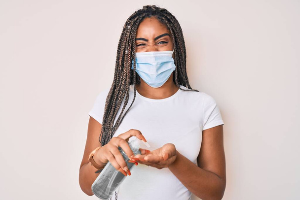 Young african american woman with braids wearing medical mask using hand sanitizer sticking tongue out happy with funny expression.  - Фото, зображення