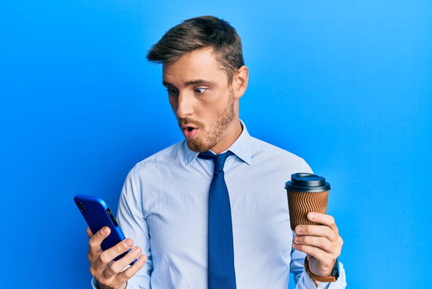 Jóképű kaukázusi üzletember használ okostelefon és iszik egy csésze kávét fél, és megdöbbent a meglepetés és csodálkozó kifejezés, félelem és izgatott arc.  - Fotó, kép