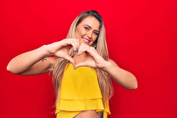 Jonge mooie blonde vrouw draagt gele t-shirt staan over geïsoleerde rode achtergrond glimlachen in liefde doen hart symbool vorm met de handen. Romantisch concept. - Foto, afbeelding