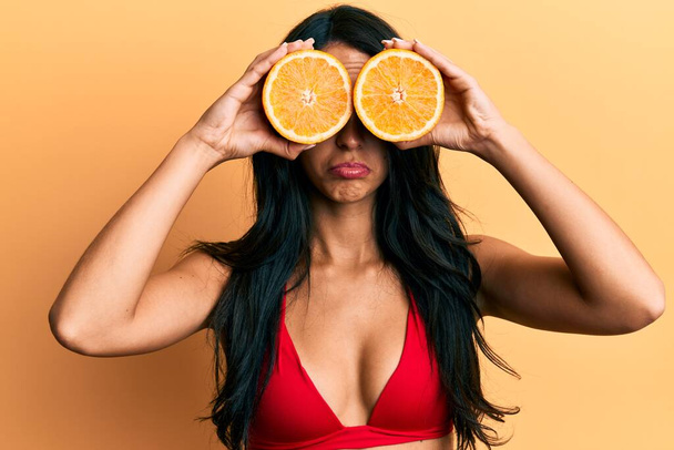 Beautiful hispanic woman wearing bikini holding orange on eyes depressed and worry for distress, crying angry and afraid. sad expression.  - 写真・画像