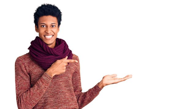 カジュアルな冬のセーターとスカーフを身に着けている若いアフリカ系アメリカ人の男性は、手で提示し、指で指している間、カメラに驚いて笑顔.  - 写真・画像
