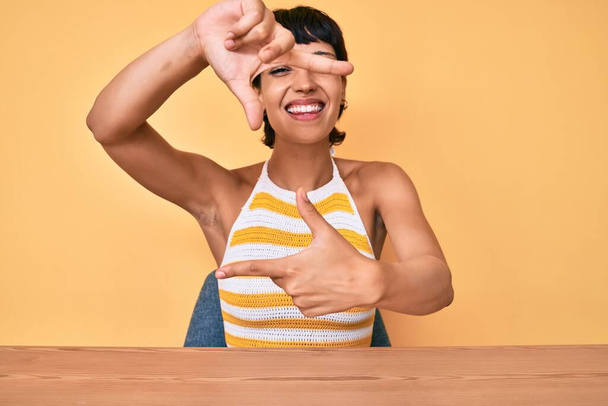 Beautiful brunettte woman sitting on the table over yellow background smiling making frame with hands and fingers with happy face. creativity and photography concept.  - Photo, Image