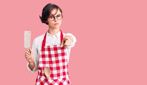 Красива молода жінка з коротким волоссям, одягнена в професійний пекарський фартух, тримає ніж, вказуючи пальцем на камеру і для вас, впевнений жест виглядає серйозно
  - Фото, зображення