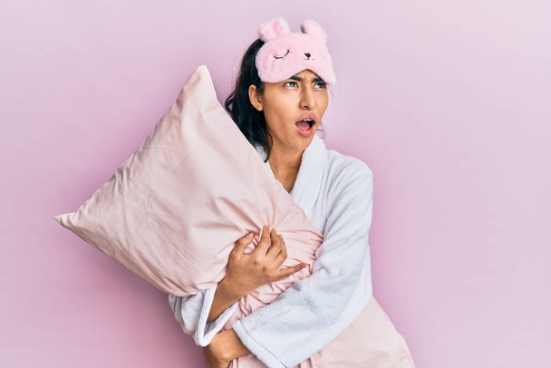 Hispanic teenager girl with dental braces wearing sleep mask and robe hugging pillow angry and mad screaming frustrated and furious, shouting with anger. rage and aggressive concept.  - Photo, Image