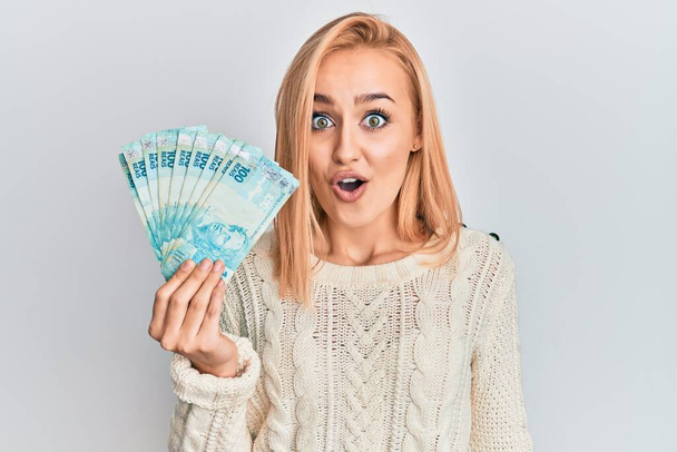Schöne kaukasische blonde Frau, die 100 echte brasilianische Banknoten in der Hand hält, erschrocken und erstaunt mit offenem Mund für Überraschung, ungläubiges Gesicht  - Foto, Bild