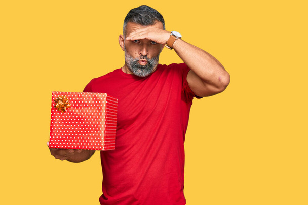 Middle age handsome man holding gift stressed and frustrated with hand on head, surprised and angry face  - Photo, Image