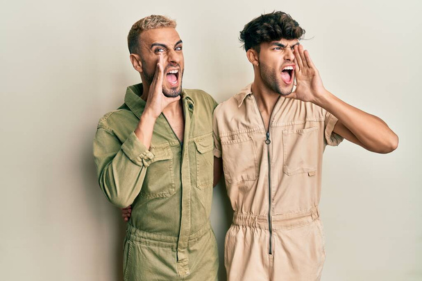 Homosexual gay couple standing together wearing casual jumpsuit shouting and screaming loud to side with hand on mouth. communication concept.  - Photo, Image