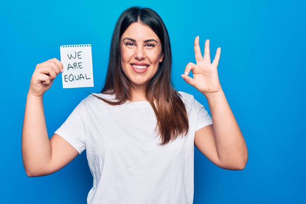 Молодая красивая брюнетка женщина просит равенства проведение бумаги с мы равны сообщение делает хорошо знак с пальцами, улыбаясь дружеский жест отличный символ - Фото, изображение