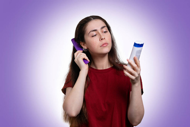 Portrait of a young woman combing her hair and reading the composition of shampoo in her hand. Purple background. Copy the space. Hair care concept. - Фото, изображение