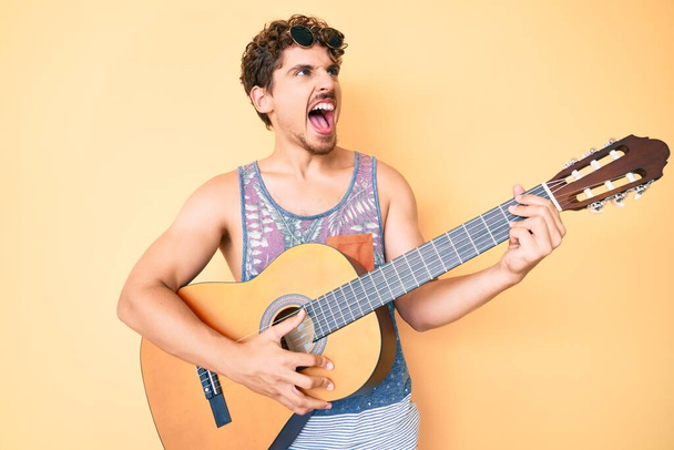 Young caucasian man with curly hair playing classical guitar angry and mad screaming frustrated and furious, shouting with anger. rage and aggressive concept.  - Photo, Image