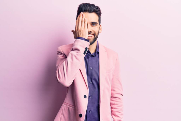 Young handsome man with beard wearing elegant jacket covering one eye with hand, confident smile on face and surprise emotion.  - Photo, Image