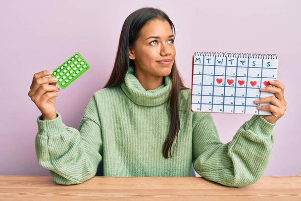 Piękna latynoska kobieta trzymająca tabletki antykoncepcyjne uśmiechająca się patrząc w bok i patrząc myśląc.  - Zdjęcie, obraz