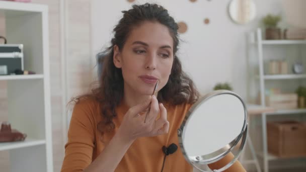 Primo piano POV del giovane vlogger di bellezza femminile caucasica che guarda nello specchio da tavolo, applica il trucco sulle labbra con il pennello, quindi rimuove l'eccesso mordendo la carta velina del viso - Filmati, video