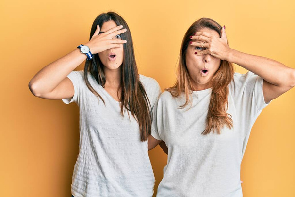Hispanic family of mother and daughter wearing casual white tshirt peeking in shock covering face and eyes with hand, looking through fingers with embarrassed expression.  - Photo, Image