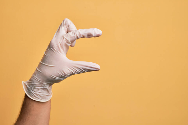 Ręka białego młodzieńca z rękawiczką medyczną nad odizolowanym żółtym tłem zbierającym i zabierającym niewidzialną rzecz, trzymającym obiekt z palcami pokazującymi przestrzeń - Zdjęcie, obraz