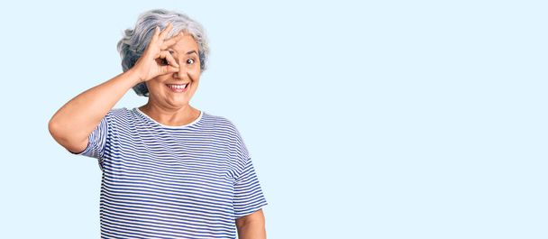 Старшая женщина с седыми волосами в повседневной полосатой одежде делает хорошо жест с улыбкой на руке, глаза смотрят сквозь пальцы с счастливым лицом.  - Фото, изображение