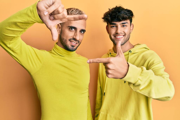 Гомосексуальна гей-пара стоїть разом у жовтому одязі, посміхаючись, роблячи рамку руками та пальцями з щасливим обличчям. креативність і концепція фотографії
.  - Фото, зображення