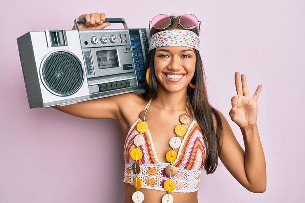 Beautiful hispanic woman wearing hippie style holding boom box doing ok sign with fingers, smiling friendly gesturing excellent symbol  - Photo, Image