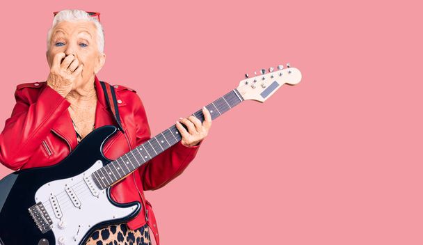Senior gyönyörű nő kék szemek és ősz haj visel modern megjelenés játszik elektromos gitár nevetés és zavarba kuncogás fedi száj kézzel, pletyka és botrány koncepció  - Fotó, kép