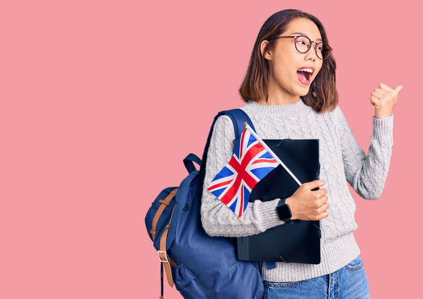 Fiatal gyönyörű kínai lány visel diák hátizsák tartó kötőanyag és Egyesült Királyság zászló mutató hüvelykujj fel az oldalra mosolyogva boldog nyitott szájjal  - Fotó, kép