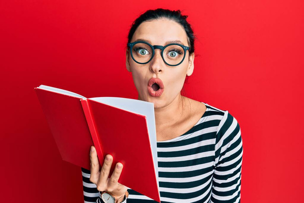 Mujer caucásica joven con gafas que cubren la boca con libro asustado y sorprendido con la boca abierta para sorpresa, cara de incredulidad  - Foto, Imagen