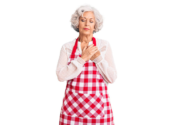 Старша сіро-волохата жінка носить фартух, посміхаючись руками на грудях із закритими очима та вдячним жестом на обличчі. концепція здоров'я
.  - Фото, зображення